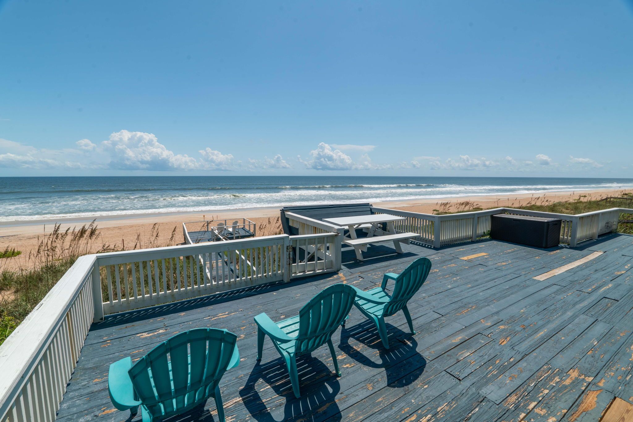 Oceanfront beach deck with weathered blue wooden floor, three turquoise Adirondack chairs, picnic table and sandy shoreline under a clear blue sky
