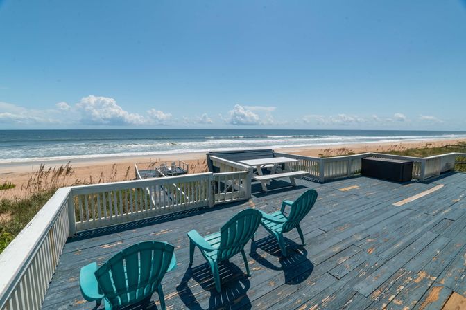Oceanfront beach deck with weathered blue wooden floor, three turquoise Adirondack chairs, picnic table and sandy shoreline under a clear blue sky