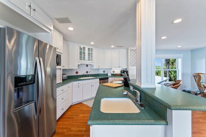 Bright coastal-style open kitchen with white cabinets, green countertops, stainless steel French-door refrigerator, breakfast bar sink, hardwood floors and palm-tree view