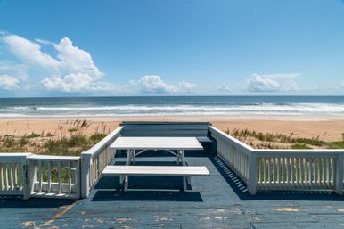 Sunny oceanfront wooden deck with a white picnic table and bench overlooking a sandy beach, gentle waves and a bright blue sky dotted with fluffy clouds.
