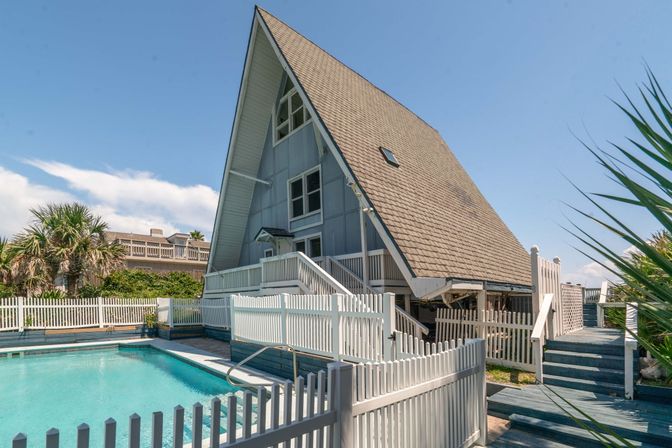 Sunlit A-frame coastal vacation home with steep shingled roof, white picket fence, palm trees and a turquoise private pool.