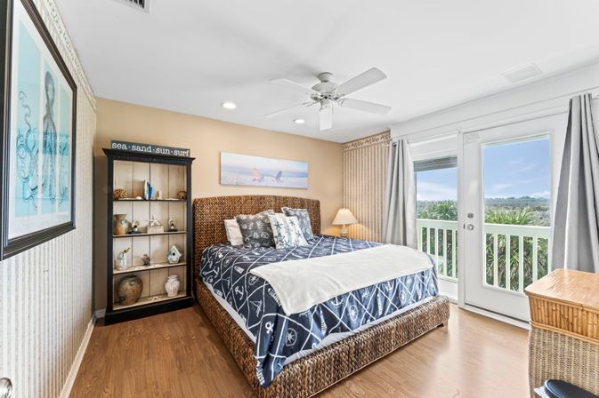 Bright coastal bedroom in a beach rental with a wicker bed, nautical bedding, a shelf sign reading 'sea-sand-sun-surf', bedside lamp, and French doors opening to a balcony with dunes and palm greenery views.