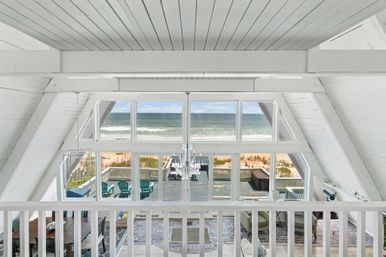 Oceanfront A-frame cottage interior with white vaulted ceiling and chandelier, floor-to-ceiling windows framing sandy beach and rolling waves, deck with teal Adirondack chairs