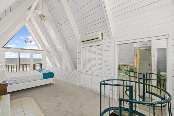 Bright A-frame coastal loft bedroom with white shiplap walls, spiral staircase and large windows opening onto an oceanfront balcony view
