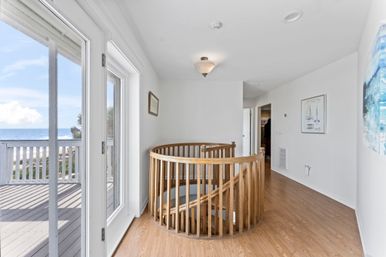 Sunny oceanfront second-floor landing in a beach house with a circular wooden spiral staircase, sliding glass doors opening to an ocean-view deck, light wood floors and white walls.