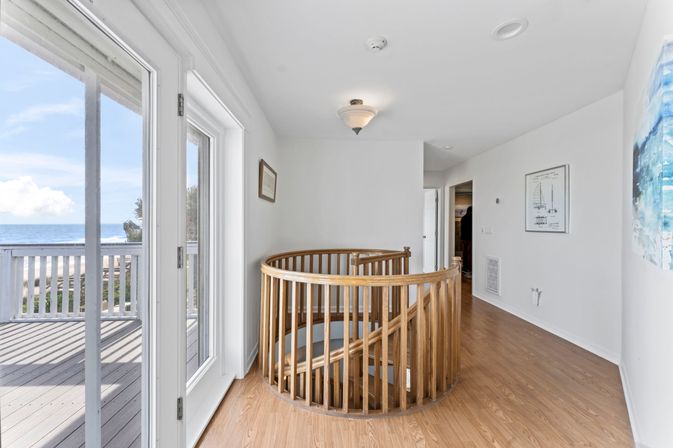Sunny oceanfront second-floor landing in a beach house with a circular wooden spiral staircase, sliding glass doors opening to an ocean-view deck, light wood floors and white walls.