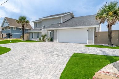 Bright coastal two-story gray house with attached two-car garage, expansive light-gray paver driveway, palm trees and neatly trimmed green lawn under a blue sky