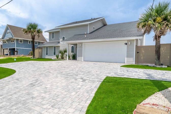 Bright coastal two-story gray house with attached two-car garage, expansive light-gray paver driveway, palm trees and neatly trimmed green lawn under a blue sky