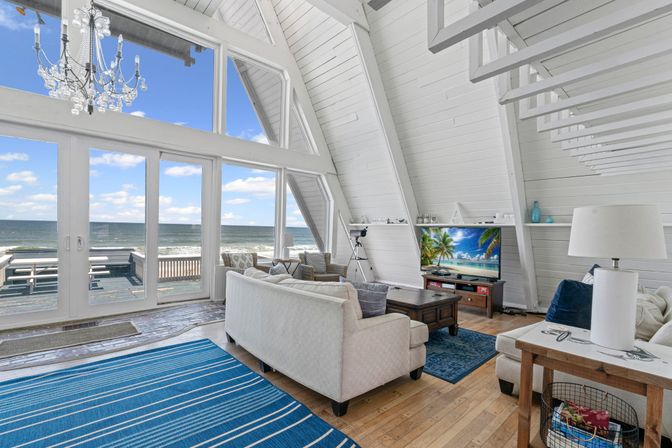 Sunlit beachfront A-frame living room with vaulted white wood ceiling, chandelier, floor-to-ceiling glass doors to oceanfront deck, cozy sofas and coastal decor.