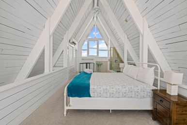 Bright A-frame attic bedroom with white shiplap beams, queen bed with teal throw, wooden nightstand, triangular window with sky view and compact en-suite sink and glass shower.