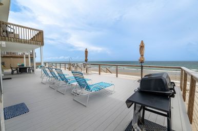 Oceanfront beach deck with row of blue-striped lounge chairs, patio table, umbrellas and a grill overlooking a sandy beach and calm sea under a cloudy sky