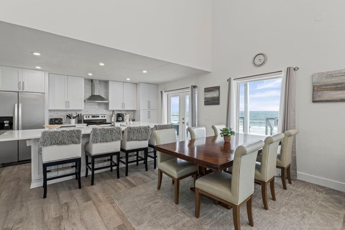 Bright open-concept beachfront kitchen and dining area with white cabinets and stainless appliances, marble island with four gray faux-fur bar stools, wooden dining table with six upholstered chairs, and sliding glass doors opening to an ocean view.