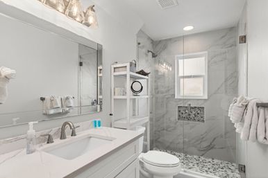 Bright modern home bathroom with white marble-look tile, frameless glass walk-in shower with pebble floor and recessed niche, white vanity with quartz countertop, brushed nickel faucet and wall mirror.