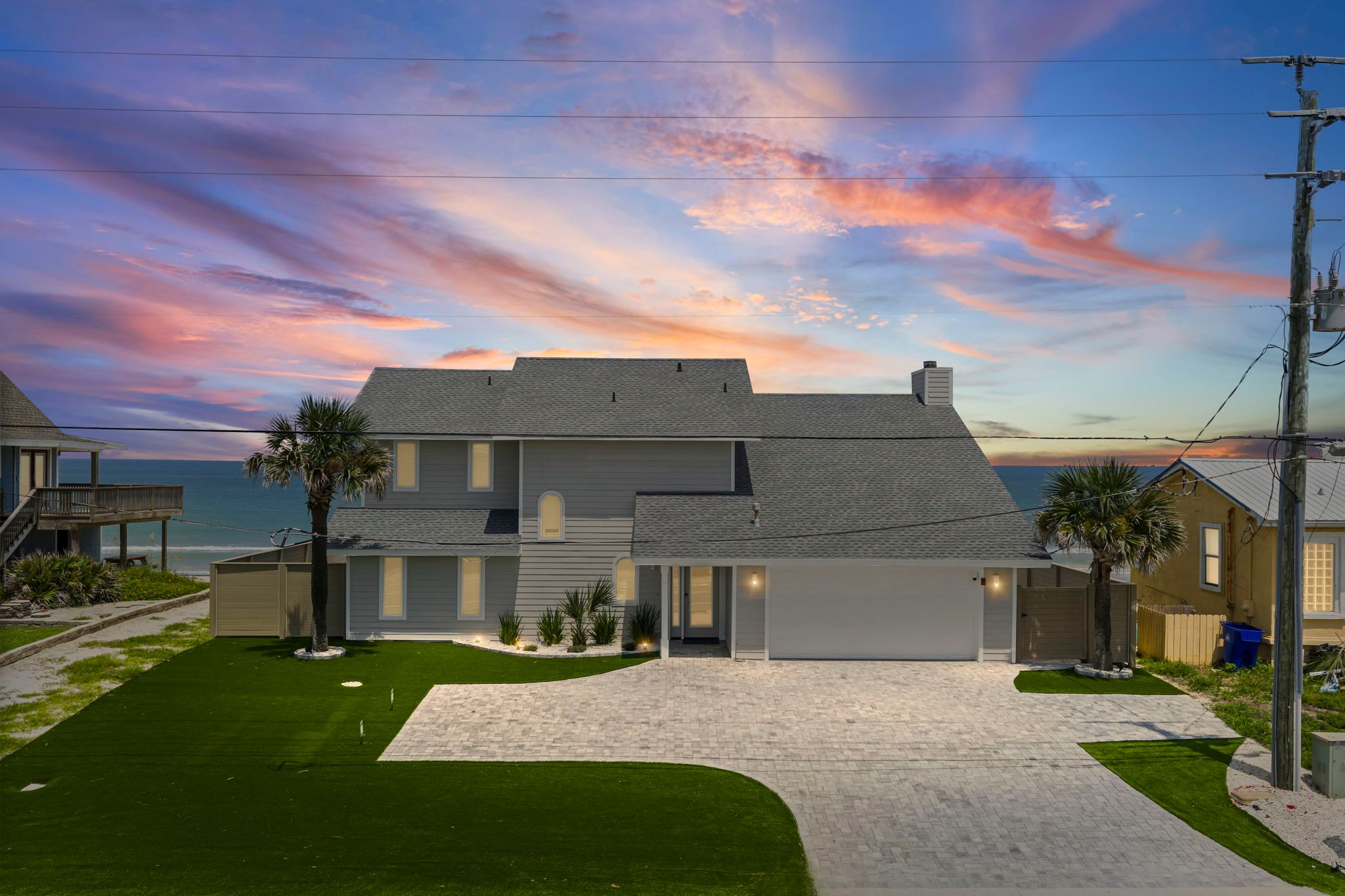 Two-story gray beachfront house with palm trees, paved driveway and manicured lawn overlooking the ocean under a colorful pink and purple sunset sky.
