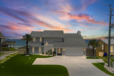 Two-story gray beachfront house with palm trees, paved driveway and manicured lawn overlooking the ocean under a colorful pink and purple sunset sky.