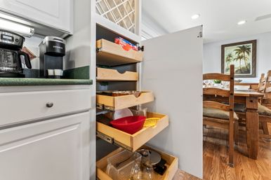 Open kitchen pull-out pantry with wooden sliding drawers holding bowls, strainers and glassware; white cabinets, green countertop with coffee makers, hardwood floor and wooden dining table with palm artwork in the background.