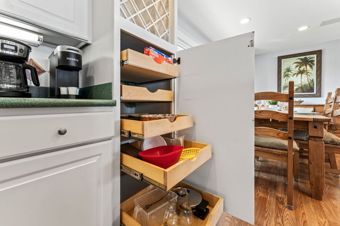 Open kitchen pull-out pantry with wooden sliding drawers holding bowls, strainers and glassware; white cabinets, green countertop with coffee makers, hardwood floor and wooden dining table with palm artwork in the background.