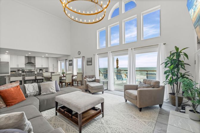 Bright beachfront open-concept living room with vaulted ceilings and arched windows, sliding glass doors to a balcony with lounge chairs and ocean views; gray sectional, armchairs, central ottoman, dining area and modern kitchen in coastal decor.