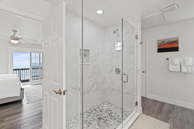Spa-like coastal en-suite with glass walk-in shower, marble tile walls and pebble floor, open door revealing ocean-view bedroom and balcony.