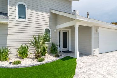 Bright modern beach-style house exterior with light gray siding, covered entry and arched windows, palm plants in white stone mulch, green lawn and paver driveway leading to attached garage.