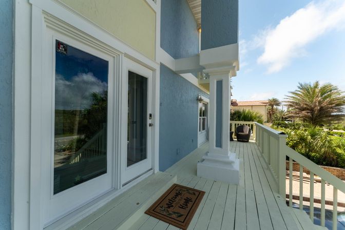 Sunny coastal balcony of a pastel beach house with white French doors and a "Welcome Home" doormat, columned railing, wicker chairs, and palm trees under a bright blue sky.