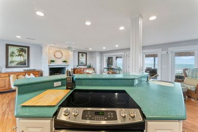 Open-concept beachfront kitchen with teal-green breakfast bar, electric cooktop, knife block and cutting board, opening to a living room with fireplace and sliding doors showing ocean views.