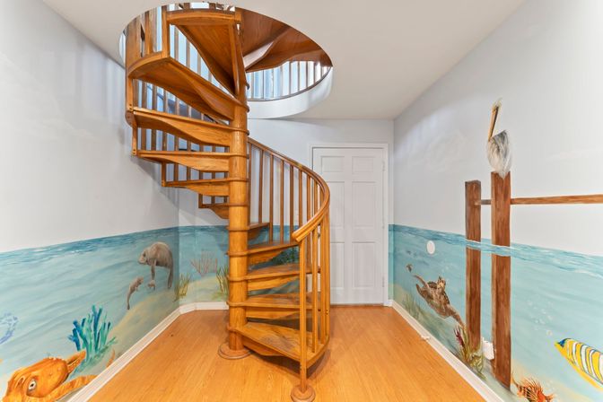 Warm oak spiral staircase rising through a circular ceiling opening in a coastal-themed interior, painted ocean mural with sea turtle, pelican on pilings, fish and coral, white door and hardwood floor.