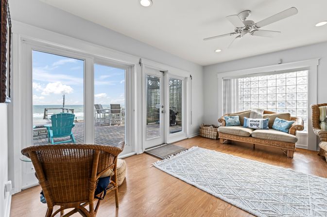 Bright beachfront living room with coastal decor — wicker sofa and chairs, hardwood floors and ceiling fan, large glass doors opening to an oceanfront patio with a turquoise Adirondack chair.