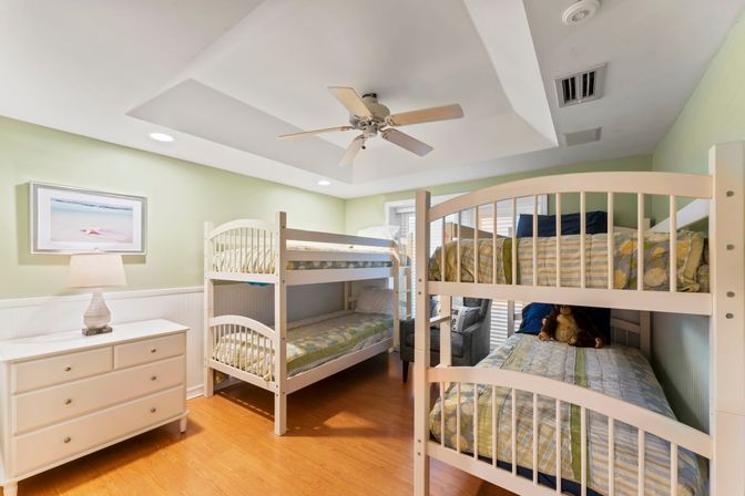 Bright coastal-style kids' bedroom with two white twin bunk beds, light green walls, hardwood floor, ceiling fan, white dresser with lamp and a framed beach print, stuffed bear on the lower bed.