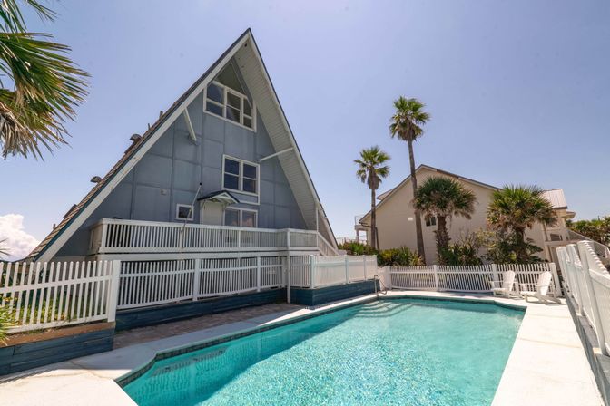 Sunlit coastal A-frame beach house with white picket fence, turquoise swimming pool in the foreground and palm trees against a clear blue sky