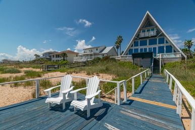 Coastal A-frame beachfront house with large windows, a blue wooden boardwalk and two white Adirondack chairs on a sandy dune under a bright blue sky