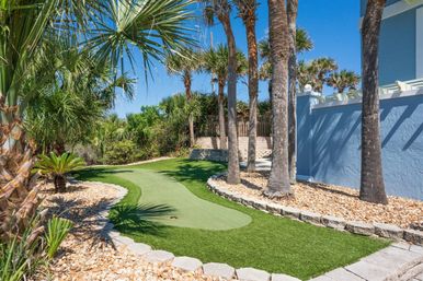 Sunny tropical coastal backyard with a curving artificial putting green flanked by palm trees, pebble beds, and a light-blue stucco wall under a clear blue sky.