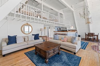 Bright coastal A-frame living room with vaulted white shiplap, mezzanine loft, two beige sofas with navy pillows, dark wood coffee table on blue rug, round porthole mirror, open kitchen and dining area.