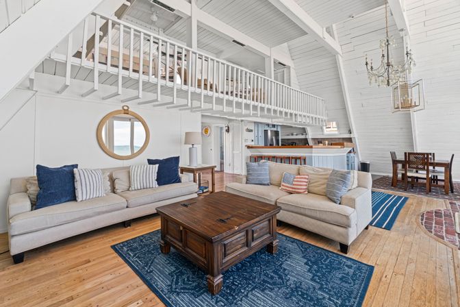 Bright coastal A-frame living room with vaulted white shiplap, mezzanine loft, two beige sofas with navy pillows, dark wood coffee table on blue rug, round porthole mirror, open kitchen and dining area.