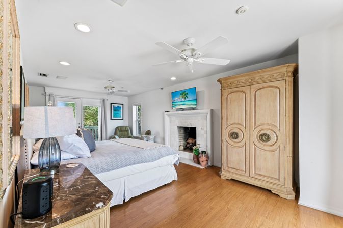 Sunlit coastal master bedroom with large bed, marble-top nightstand and lamp, ceiling fans, fireplace with wall-mounted TV, carved wood armoire, hardwood floors and balcony doors framing palm trees.