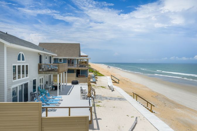 Beachfront vacation homes with elevated wooden decks and blue lounge chairs overlooking a long sandy shoreline and calm ocean under a bright blue sky