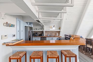 Bright beachy A-frame kitchen with white shiplap walls and exposed beams, polished wooden breakfast bar with three woven-seat stools, stainless steel refrigerator, light-blue cabinets, coffee station and adjacent dining area.