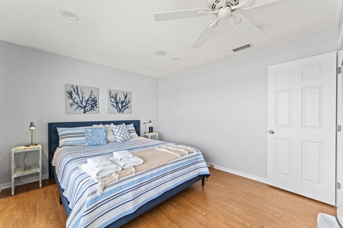 Bright coastal-style bedroom with navy upholstered bed and blue-striped linens, coral wall art above the headboard, hardwood floors, white ceiling fan, matching nightstands with lamps and neatly folded towels