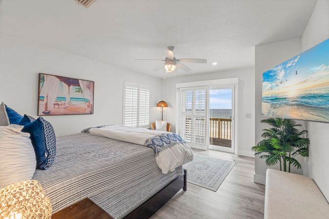 Bright beachfront master bedroom with king bed, blue coastal accents, white plantation shutters and sliding doors opening to a balcony with ocean views, ceiling fan, beach artwork and tropical plant