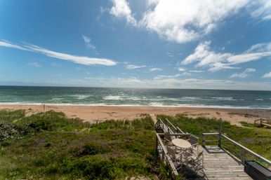 Sunny oceanfront scene showing a sandy beach and rolling waves under a blue sky, viewed from a wooden deck with a round table and chairs and a boardwalk path to the shore.
