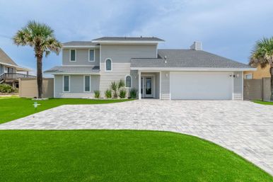Modern two-story coastal beach house with gray siding, arched windows, two-car garage, wide paver driveway, palm trees and manicured green lawn under a blue sky