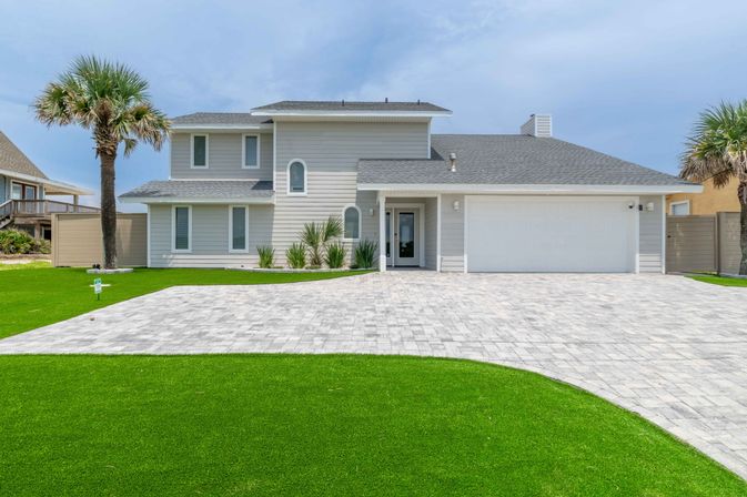 Modern two-story coastal beach house with gray siding, arched windows, two-car garage, wide paver driveway, palm trees and manicured green lawn under a blue sky