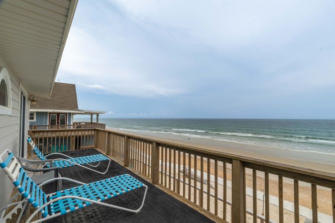 Oceanfront beach-house balcony with turquoise woven lounge chairs and wooden railing overlooking a sandy shoreline and gentle waves under a cloudy sky.