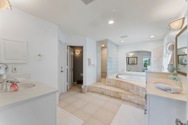 Bright airy master bathroom with raised tiled soaking tub, glass-block walk-in shower, twin vanities and light beige floor tiles