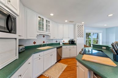 Bright coastal kitchen with white cabinets, green countertops, stainless appliances, hardwood floors, center island cooktop and cutting board, built-in wine rack and breakfast bar with stools, window framing palm trees and water view.