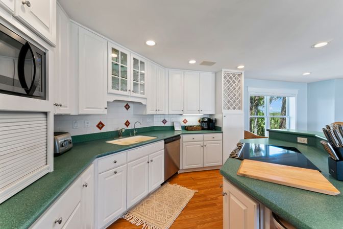 Bright coastal kitchen with white cabinets, green countertops, stainless appliances, hardwood floors, center island cooktop and cutting board, built-in wine rack and breakfast bar with stools, window framing palm trees and water view.