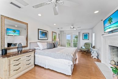 Bright coastal master bedroom with king bed, hardwood floors, white ceiling fans, marble-top dresser, fireplace, wall-mounted TV, green lounge chair and balcony with water view.