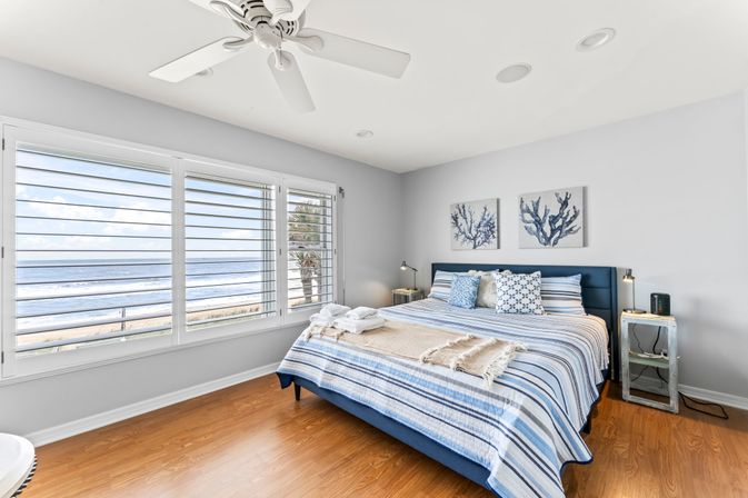 Sunlit coastal bedroom with king bed in blue-striped linens, coral wall art, hardwood floors and large shuttered windows with ocean view - beachy, relaxed vibe.