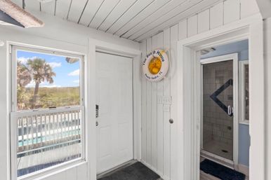 Sunny coastal entryway with white wood-paneled walls, a front door and window framing palm trees and dunes, a decorative life buoy on the wall, and an open doorway to a tiled shower.