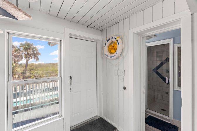 Sunny coastal entryway with white wood-paneled walls, a front door and window framing palm trees and dunes, a decorative life buoy on the wall, and an open doorway to a tiled shower.